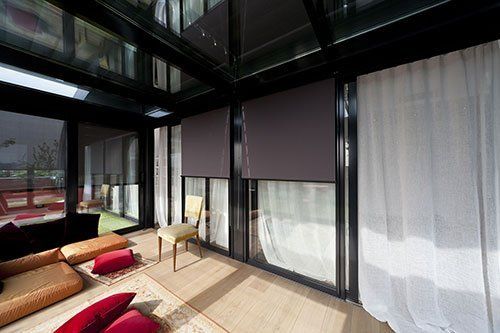 una veranda con una sedia, dei cuscini su un  pavimento in legno e delle vetrate con le tende di color bianco