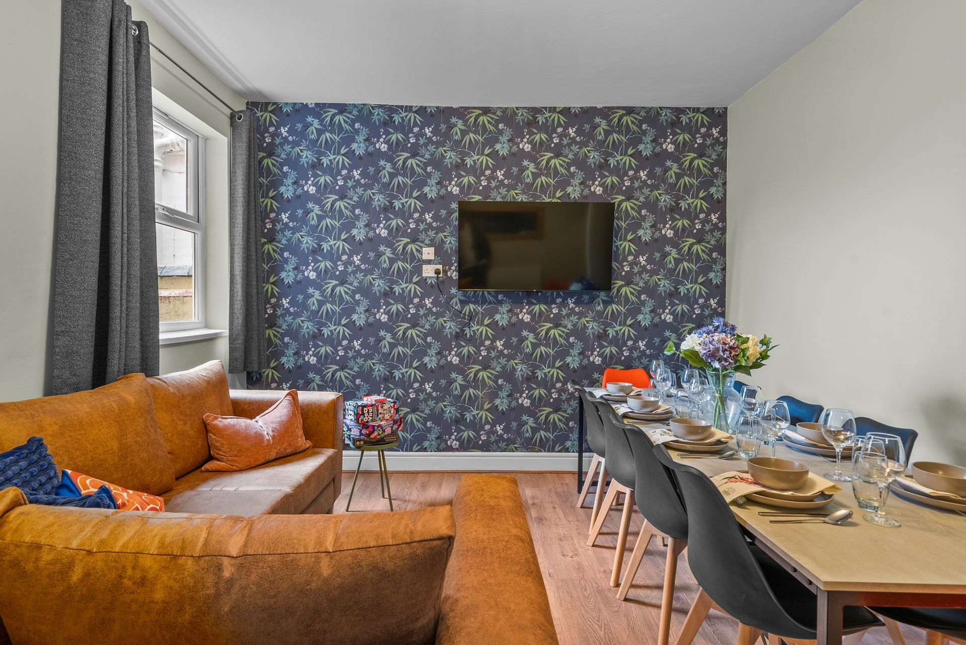Living room with floral accent wall, wall-mounted TV, orange sofa, and dining table set for a meal