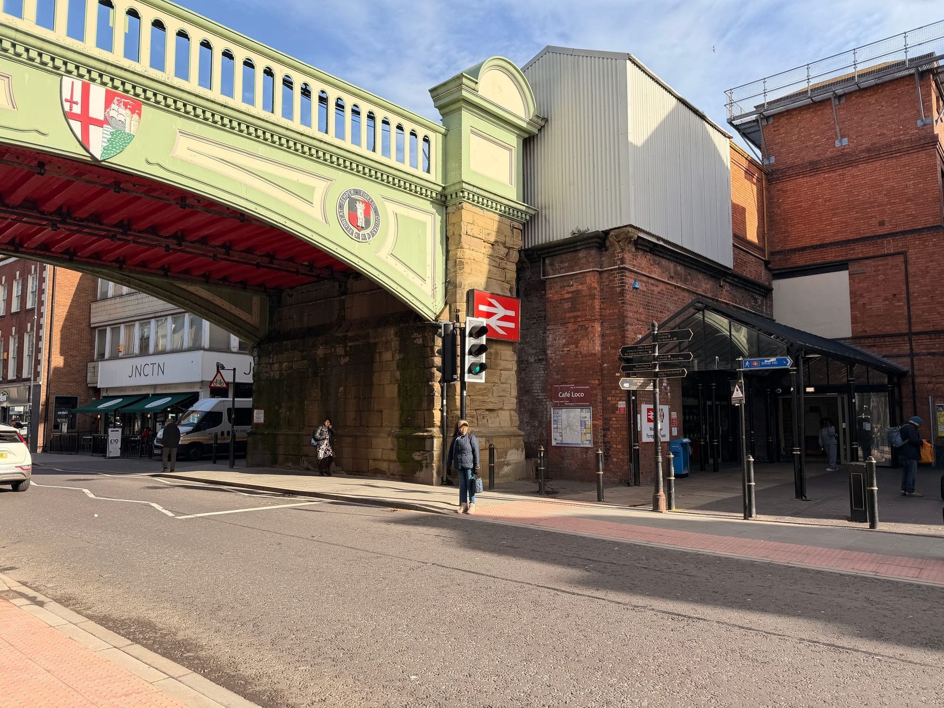 Street view of a green-painted railway bridge over a brick building and sidewalk storefronts on a sunny day