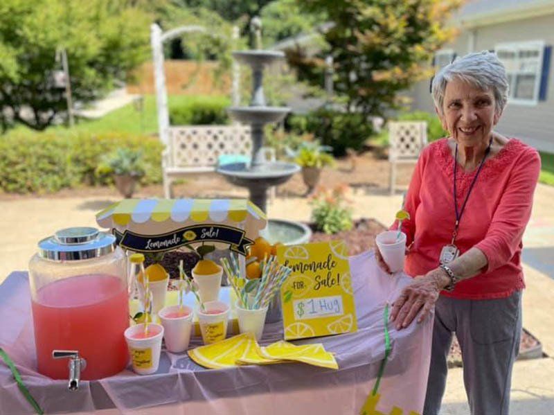 An elderly woman is standing in front of a lemonade stand.