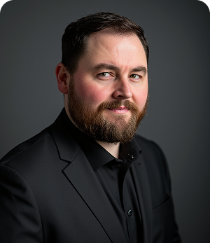 A man with a beard is wearing a black suit and black shirt.