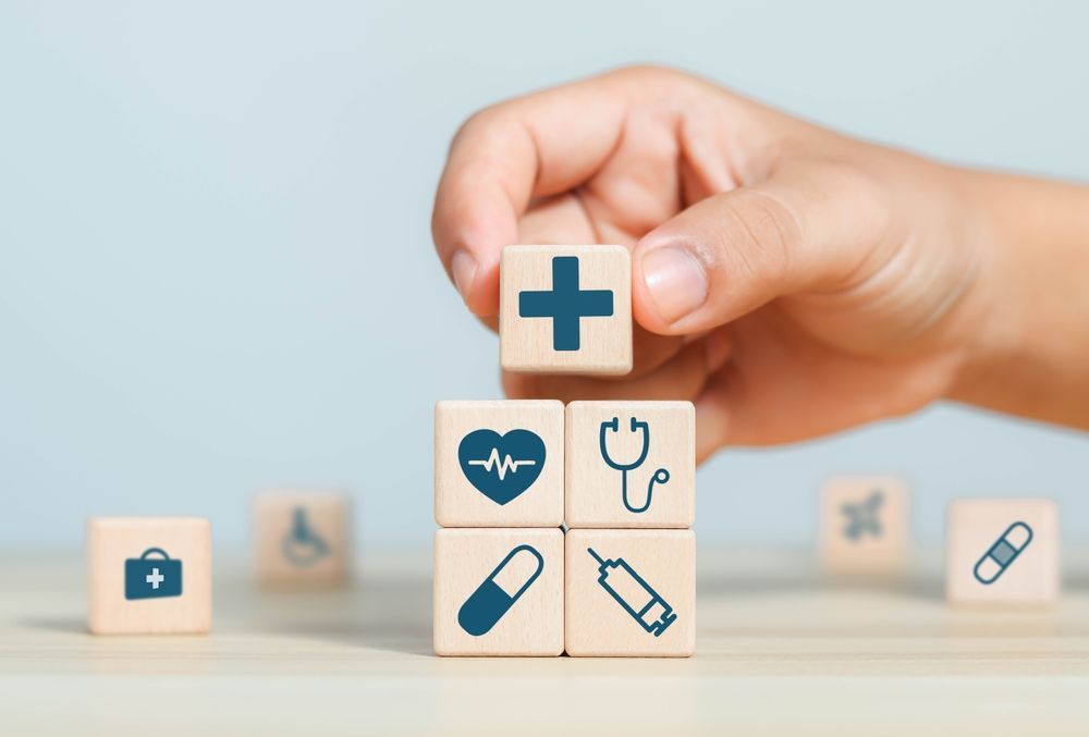 Hand placing a blue medical cross block on a stack of healthcare icons on wooden cubes