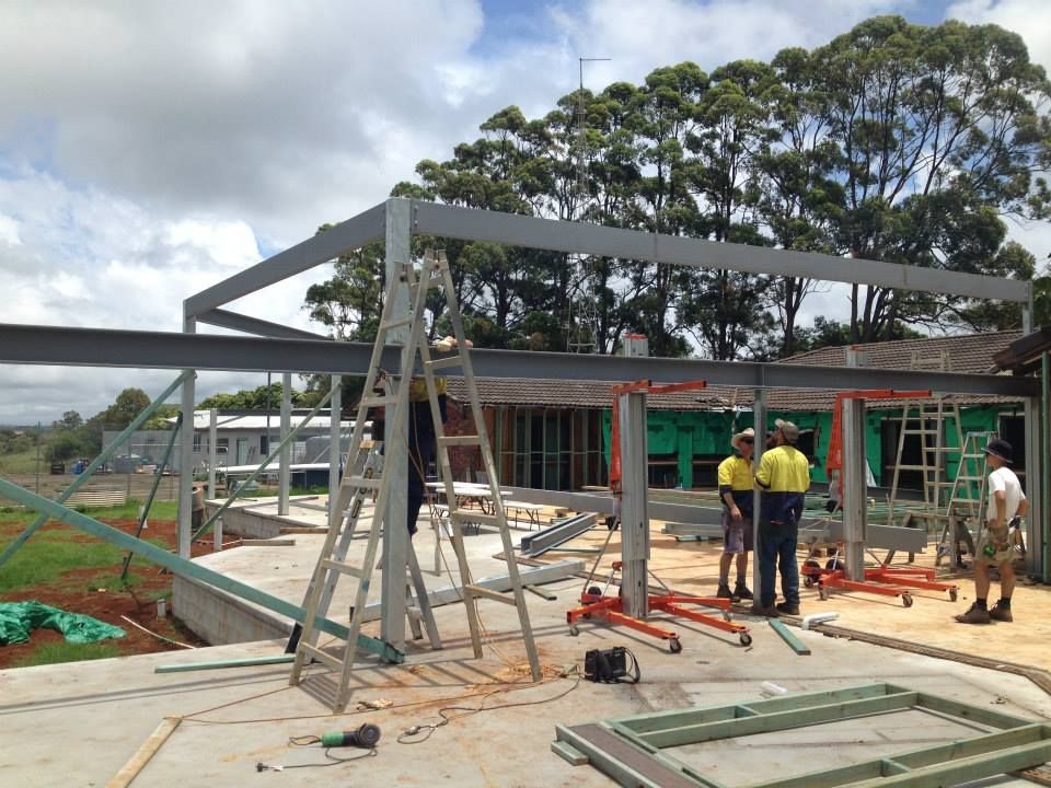 Construction site with steel frame structure; workers in yellow shirts, ladder, tools. Outdoors, overcast - North Coast Steel Fabrication in South Lismore, NSW