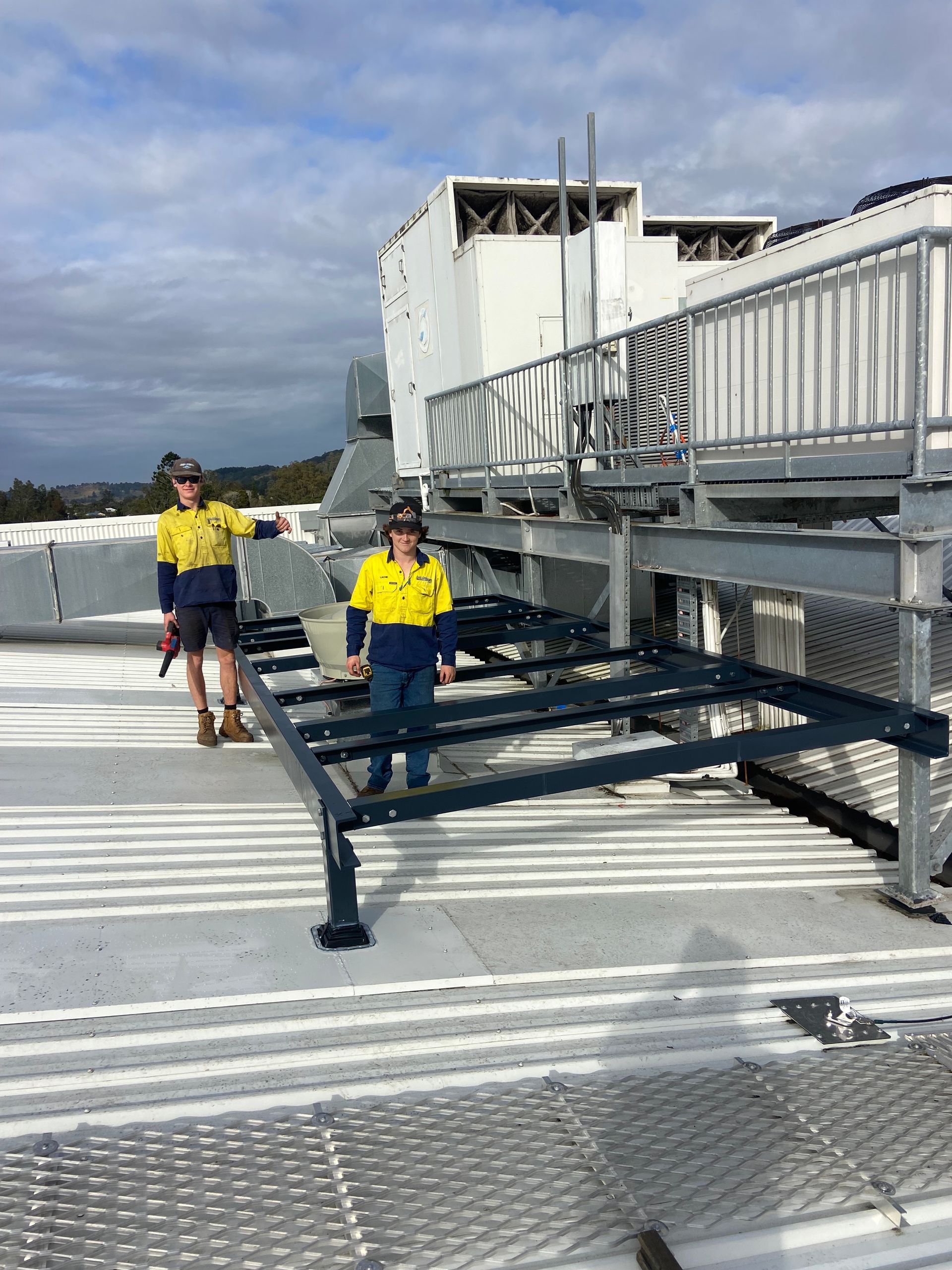 Two workers in safety gear on a rooftop, near a metal platform and equipment. Blue skies.