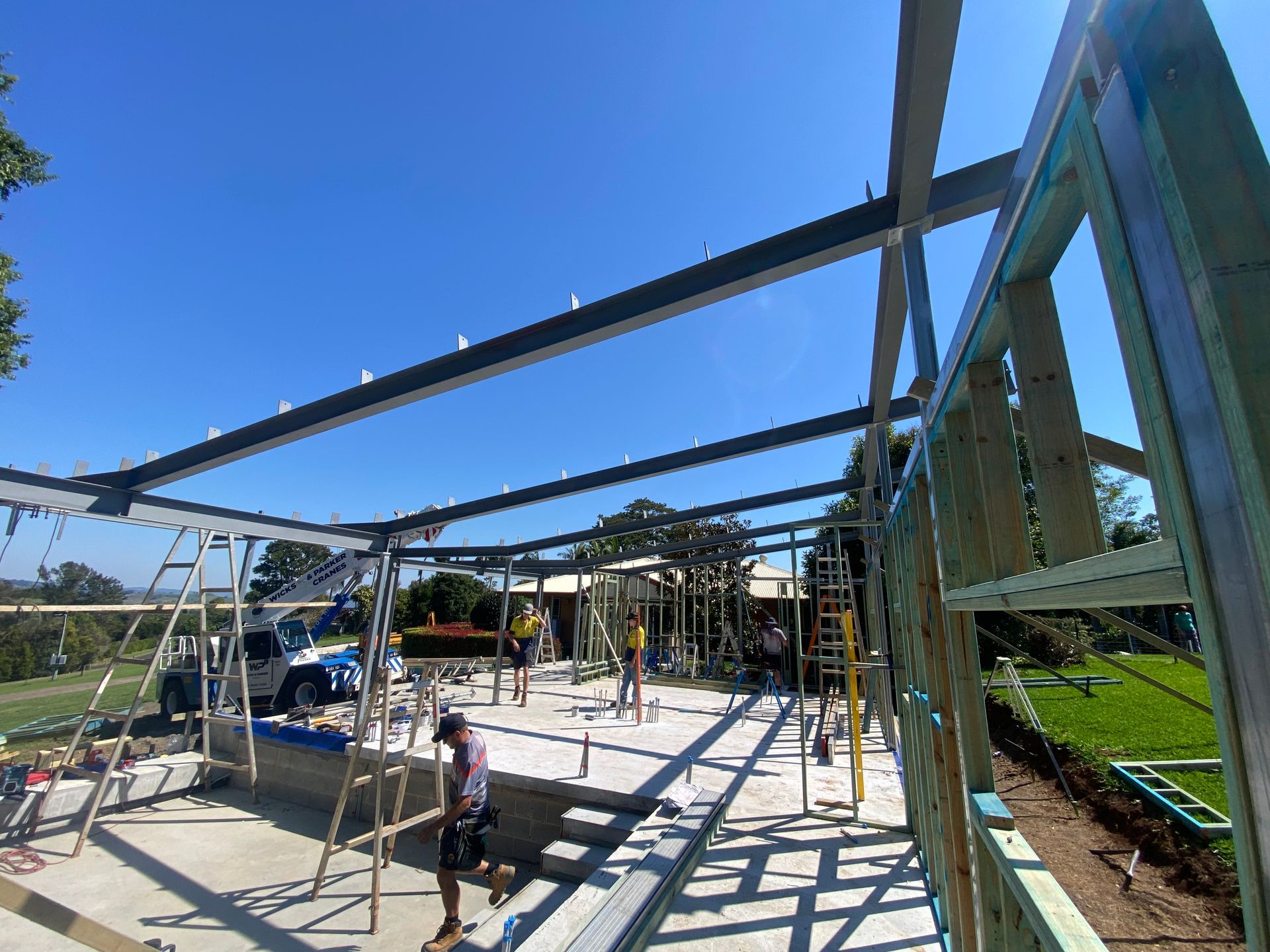 Construction site with steel beams and wooden framing under a blue sky, workers visible.