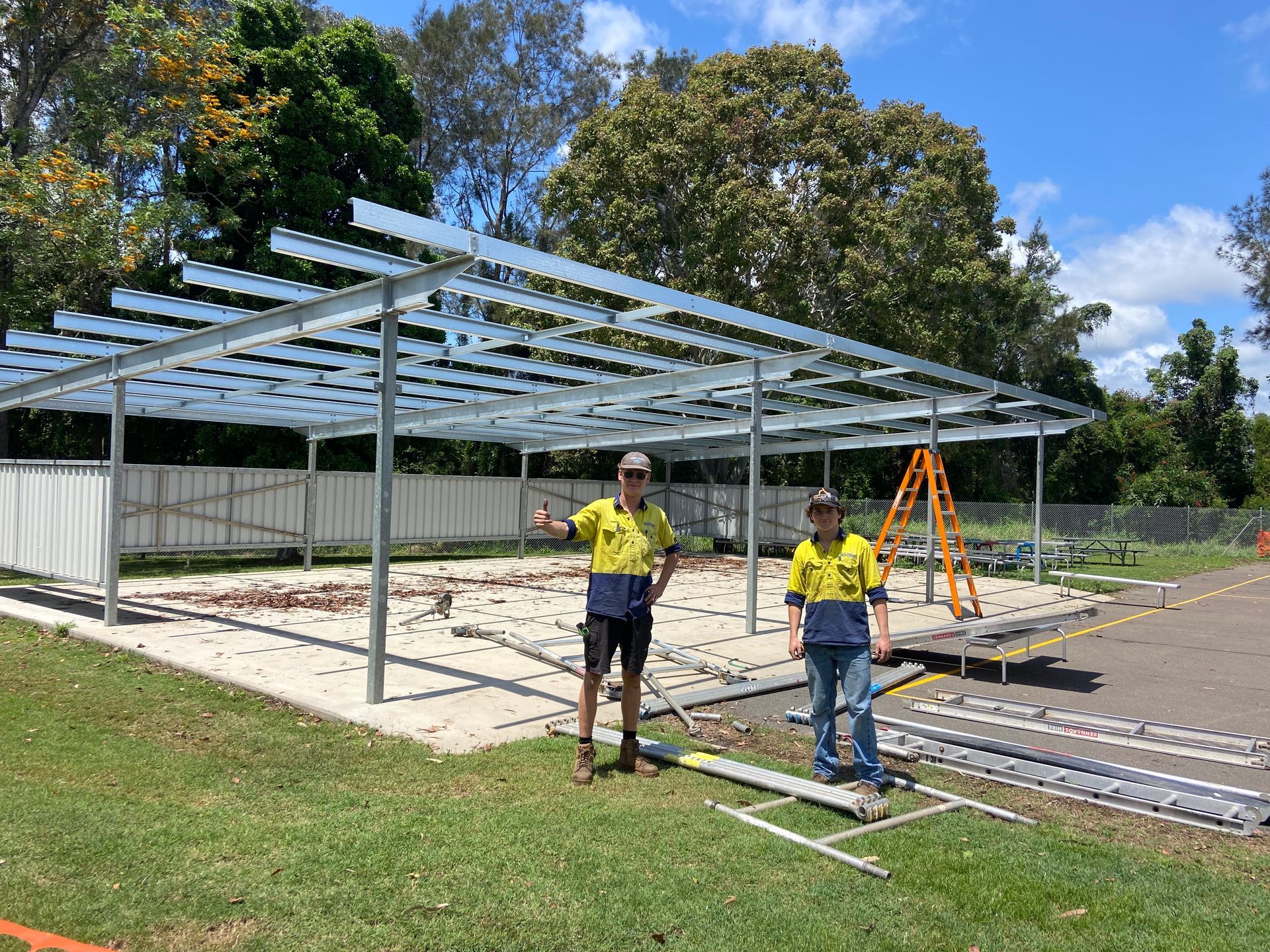 Two people stand under a partially constructed metal shelter in a park.