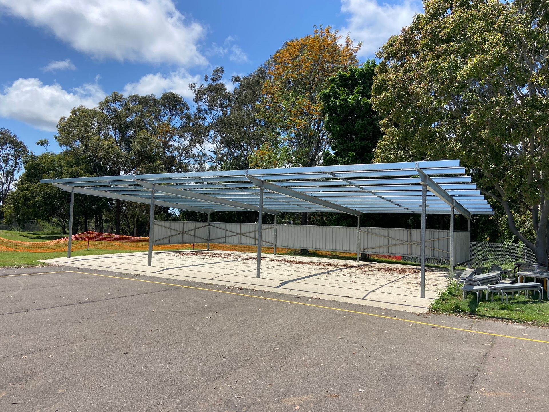 Metal carport with solar panels on a concrete base, trees and cloudy blue sky in the background.