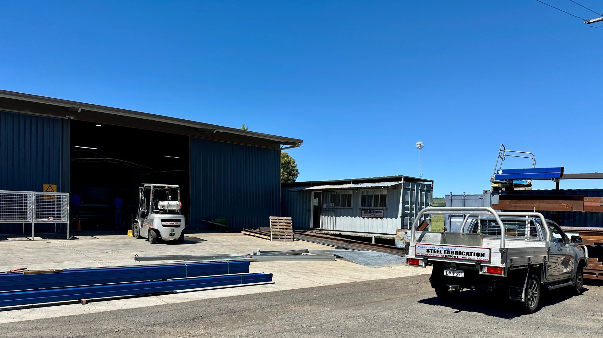 Forklift and pickup truck outside a blue industrial building on a sunny day.