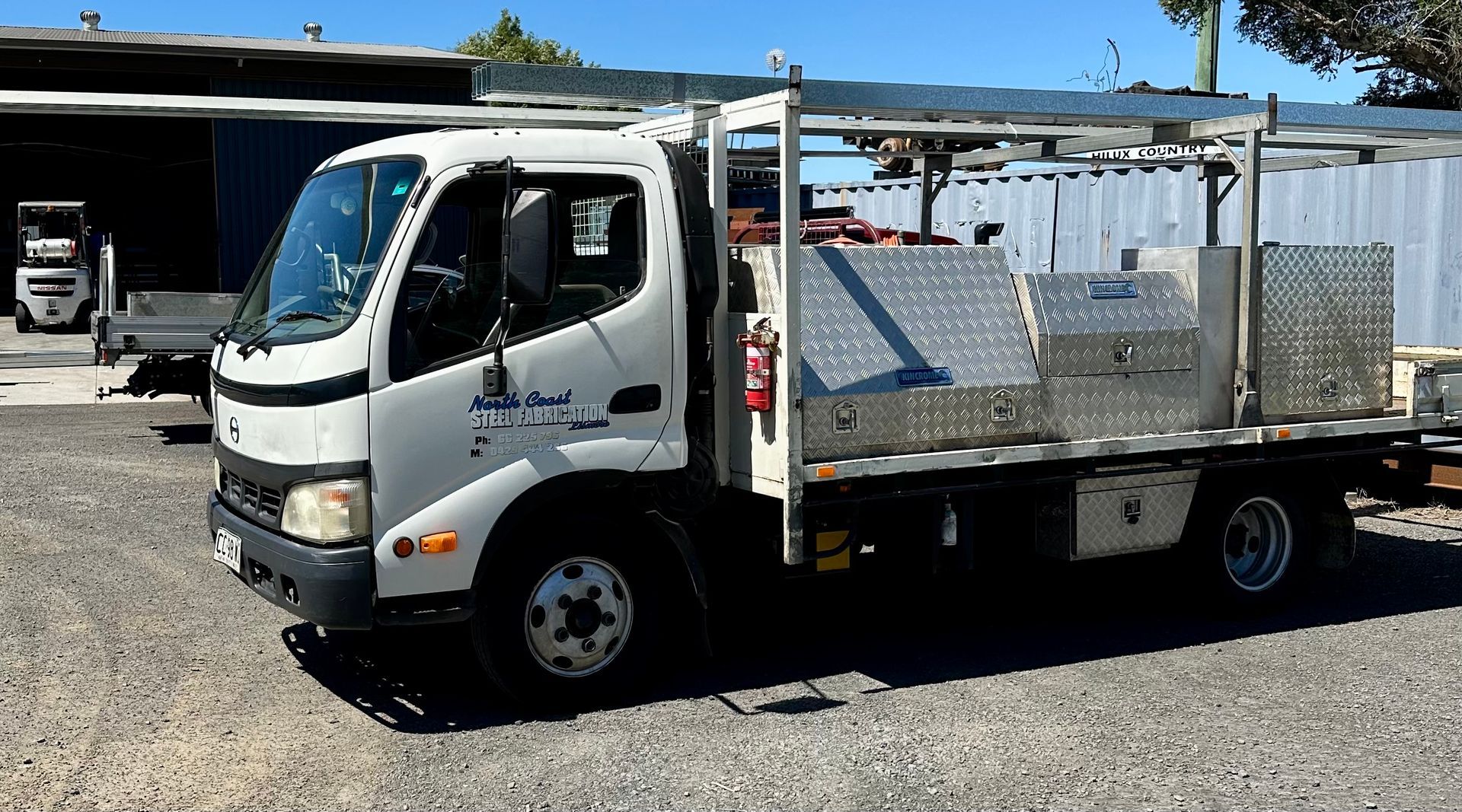 White utility truck with metal storage boxes, parked in front of a building on a gravel surface.