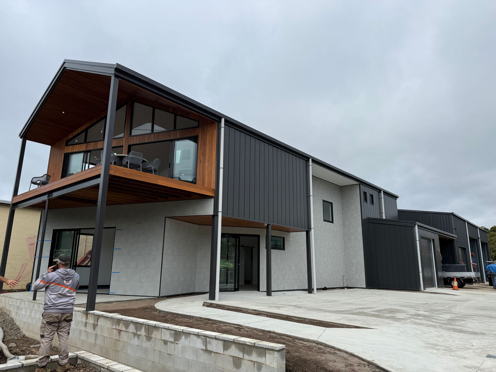 Modern two-story house with dark siding and wood accents, balcony, and a person walking in the foreground.
