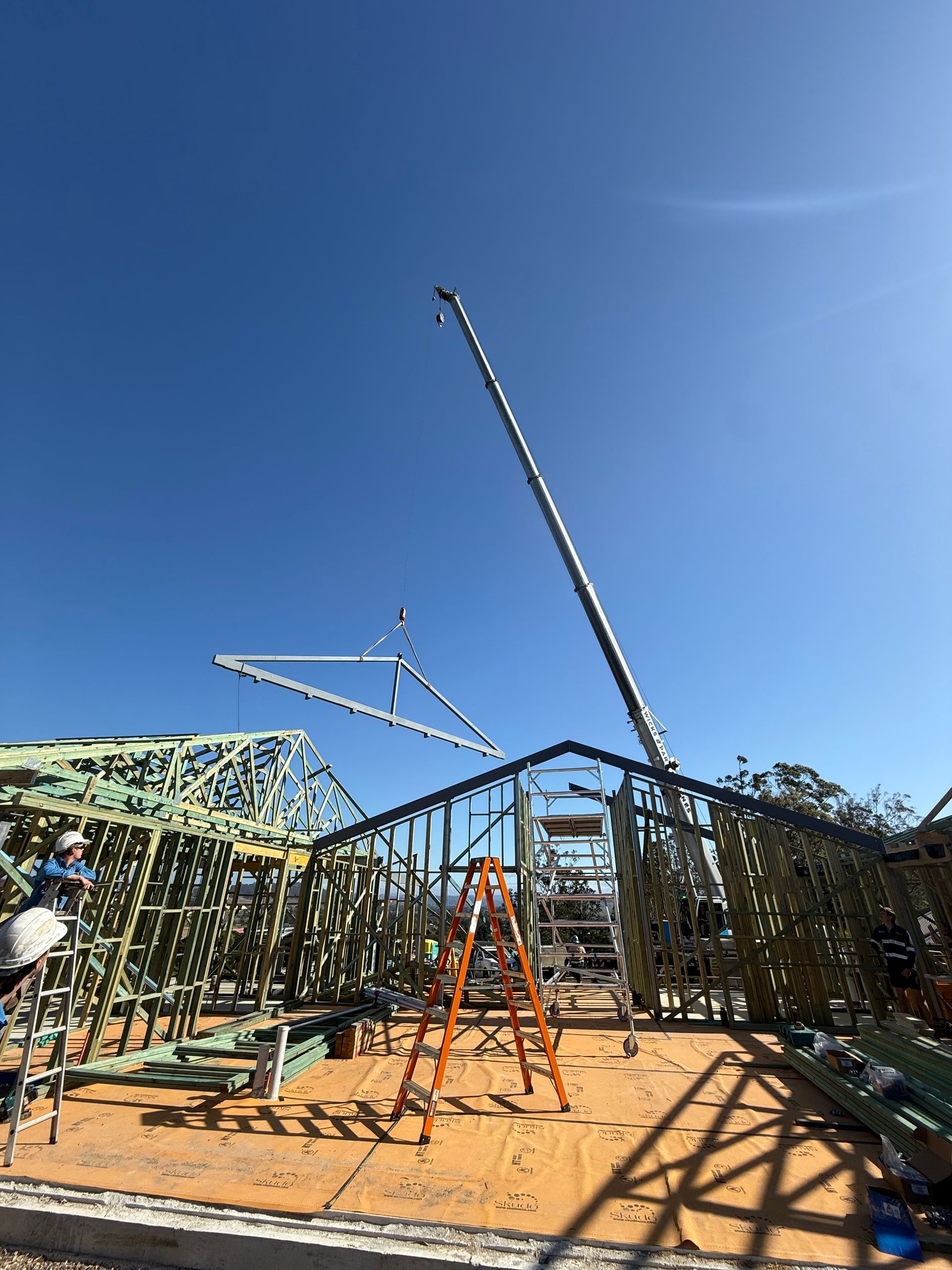 Construction site: crane lifting a roof truss onto a wooden house frame under a clear, blue sky.