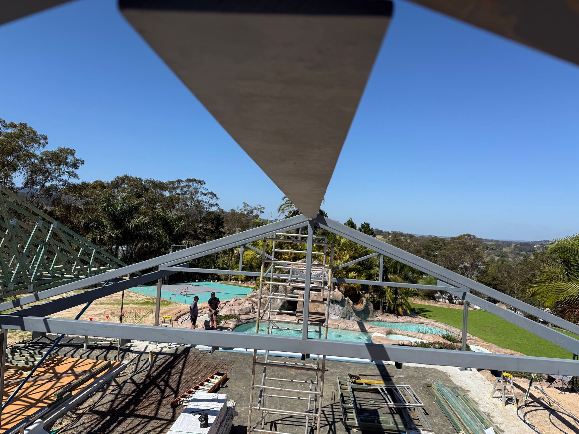 Steel frame construction over pool area with workers, trees, and blue sky.