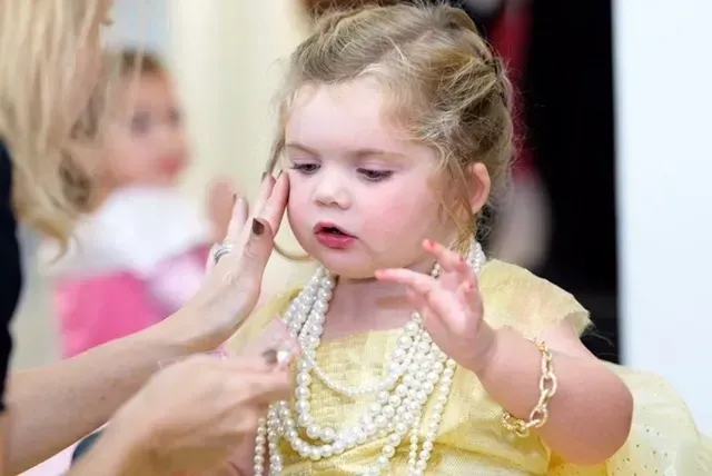 Young child in yellow dress with pearls; someone adjusts her jewelry.