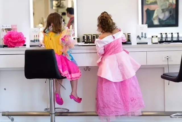 Two young children in princess costumes at a makeup counter. One sits, the other stands, both facing the counter.