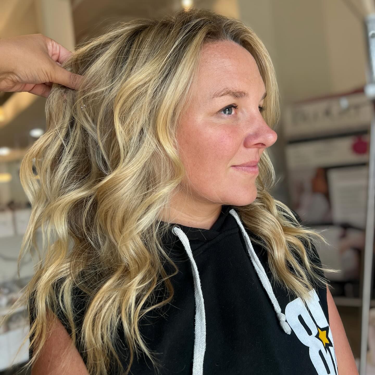 Woman with blonde wavy hair, wearing a black hoodie, hair styled, lit indoors.