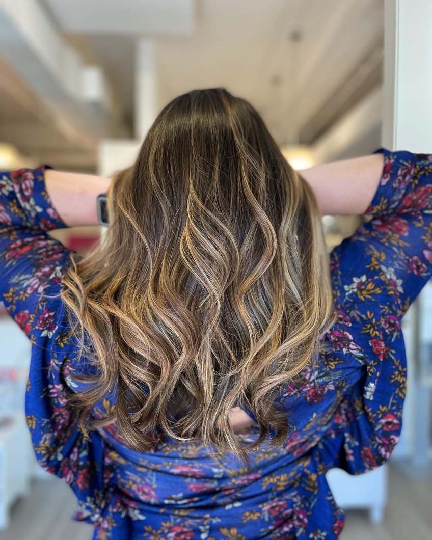 Woman with wavy, highlighted brown hair, wearing a blue floral patterned shirt, hands behind head.