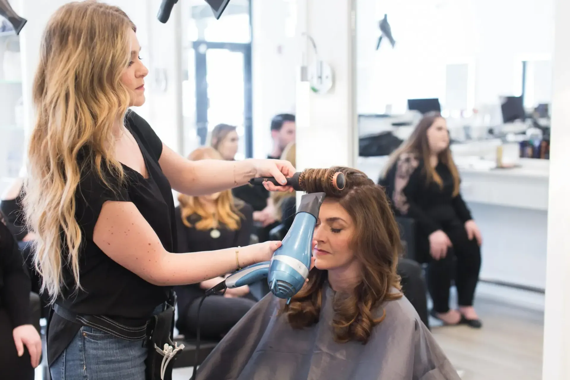 Hair stylist blow-drying client's hair in salon, using a blue hair dryer. Several people are visible in background.