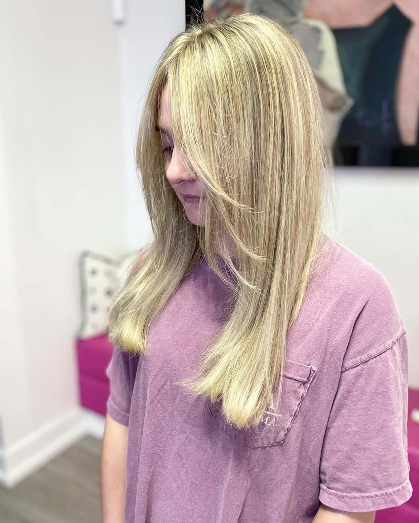 Woman with blonde hair wearing a purple shirt, posing in front of a pink bench.