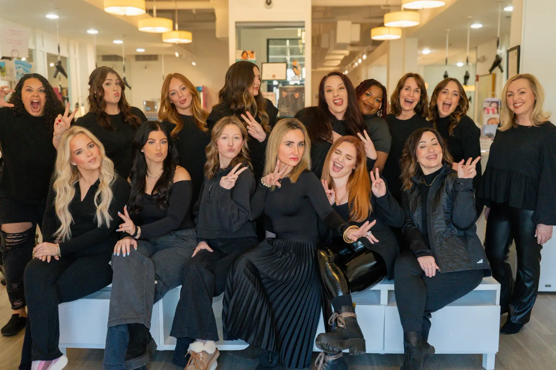 Group of women in a brightly lit salon, all in black, making playful hand gestures at the camera.