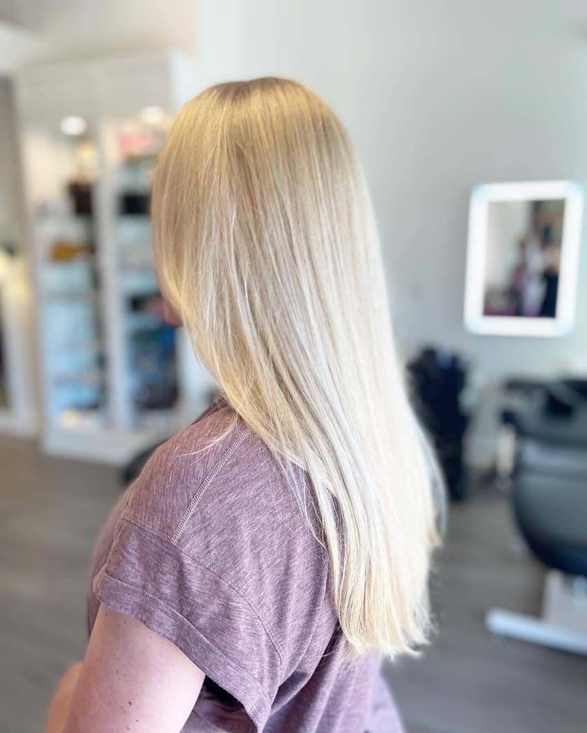 Woman with long, blonde hair; standing in a salon with a light purple shirt.