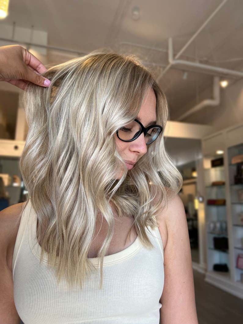 Woman with wavy blonde hair, wearing glasses and a cream top, inside a salon.