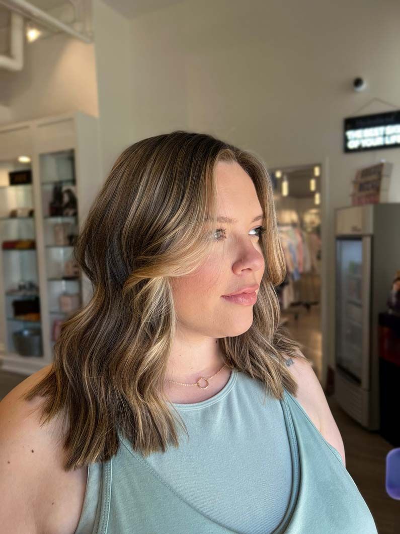 Woman with wavy brown hair, highlights, and a teal tank top in a salon.
