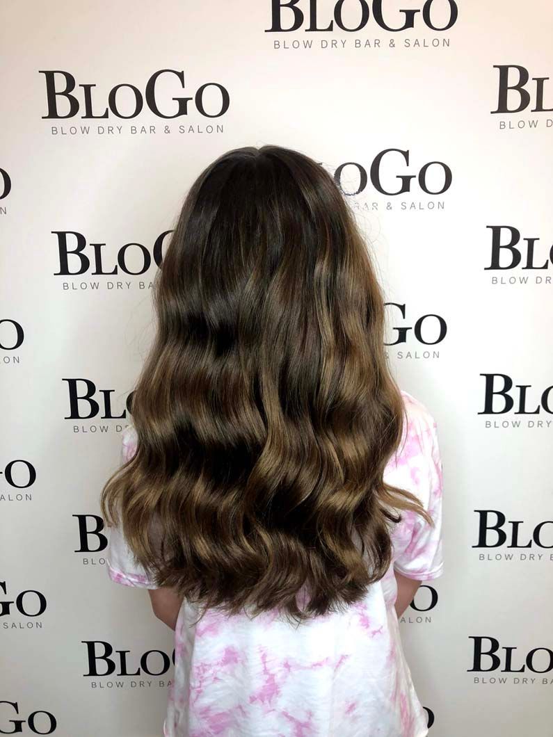 Young person with long, wavy brown hair, wearing a tie-dye shirt, standing in front of a salon logo background.