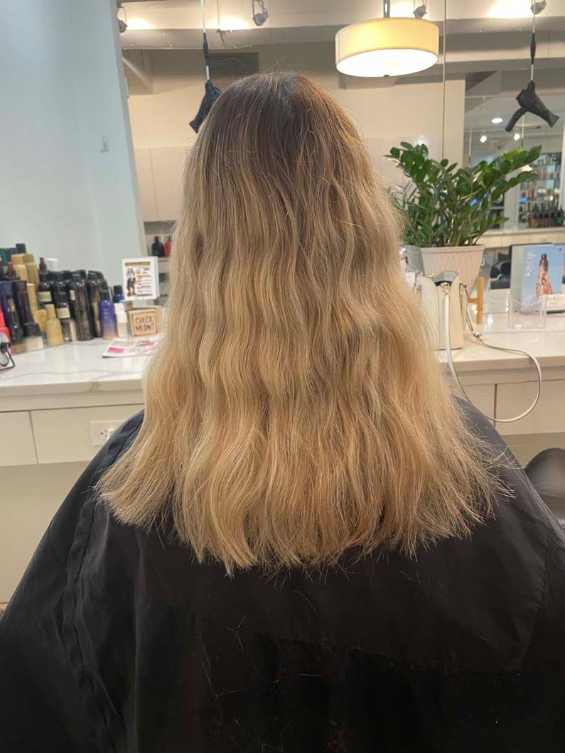 Woman with long, wavy blonde hair at a salon. Dark roots fade to a lighter shade.