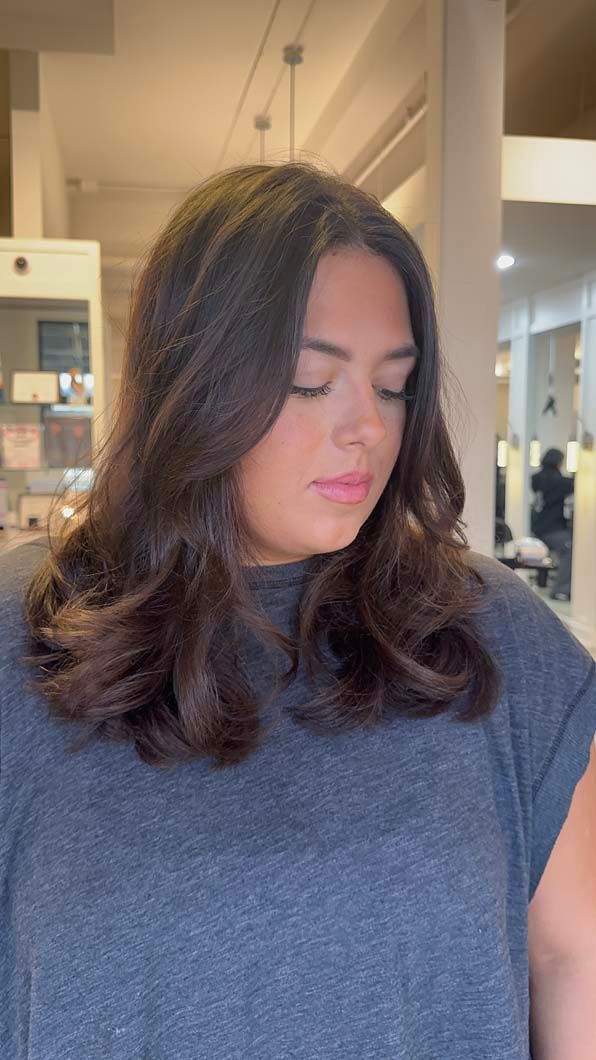 Woman with dark wavy hair looking down, wearing a gray shirt, in a salon.