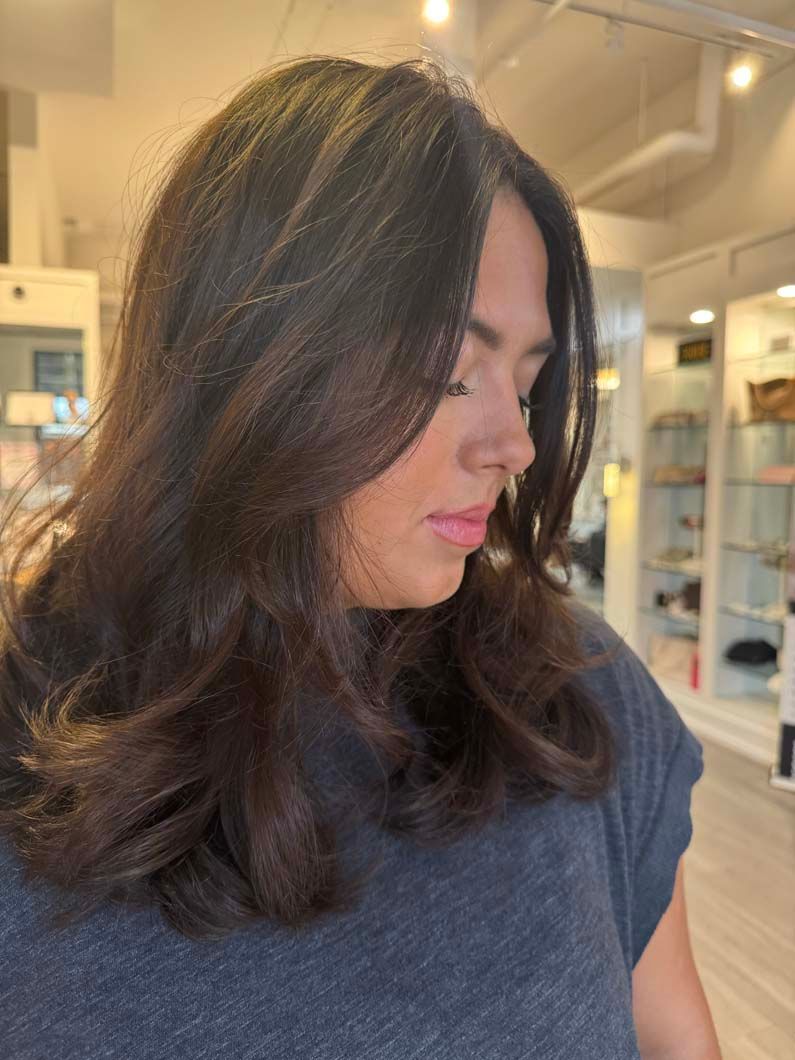 Woman with dark brown hair, looking down with a slight smile. Inside a salon setting.