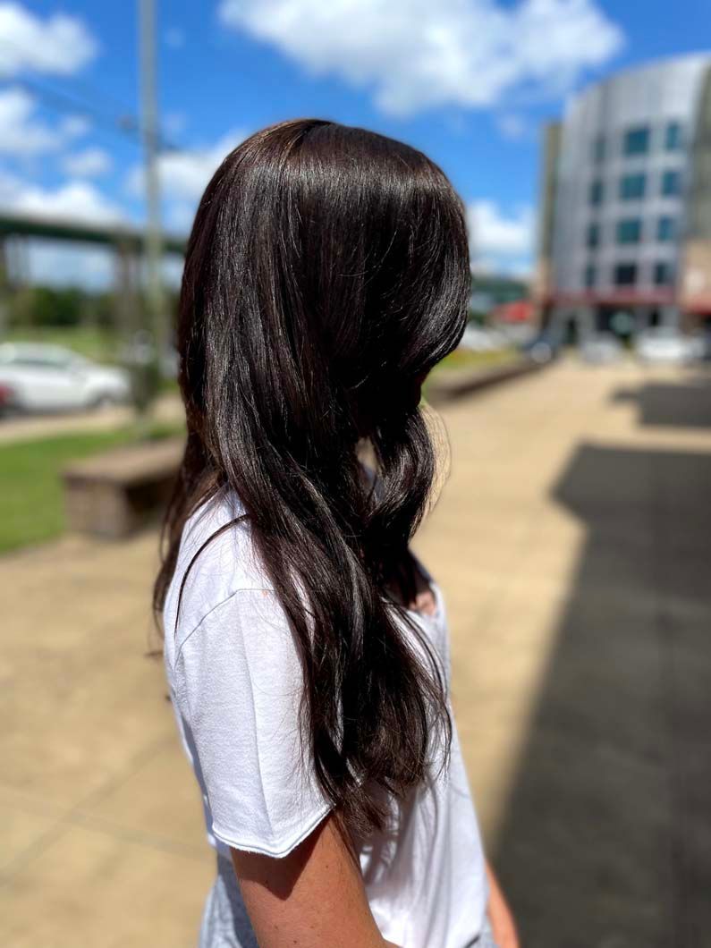 Woman with long, shiny, dark hair; wearing a white t-shirt; outdoors in sunlight.