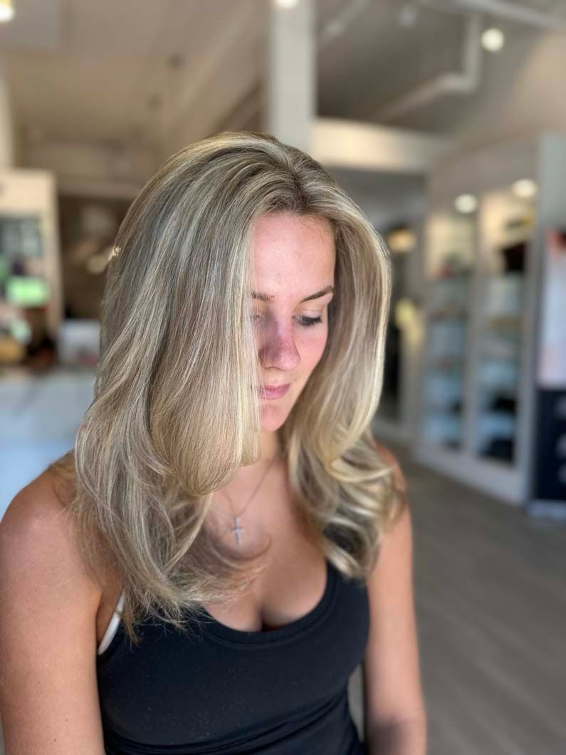 Woman with layered blonde hair in a salon, looking down, wearing a black tank top.