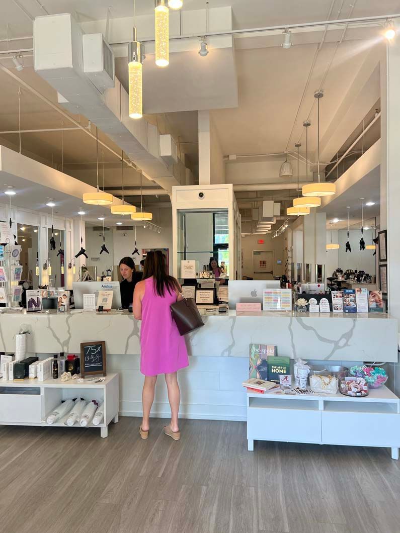 Woman in pink dress at a store counter, looking at a clerk. White walls, wood floor, merchandise displayed.