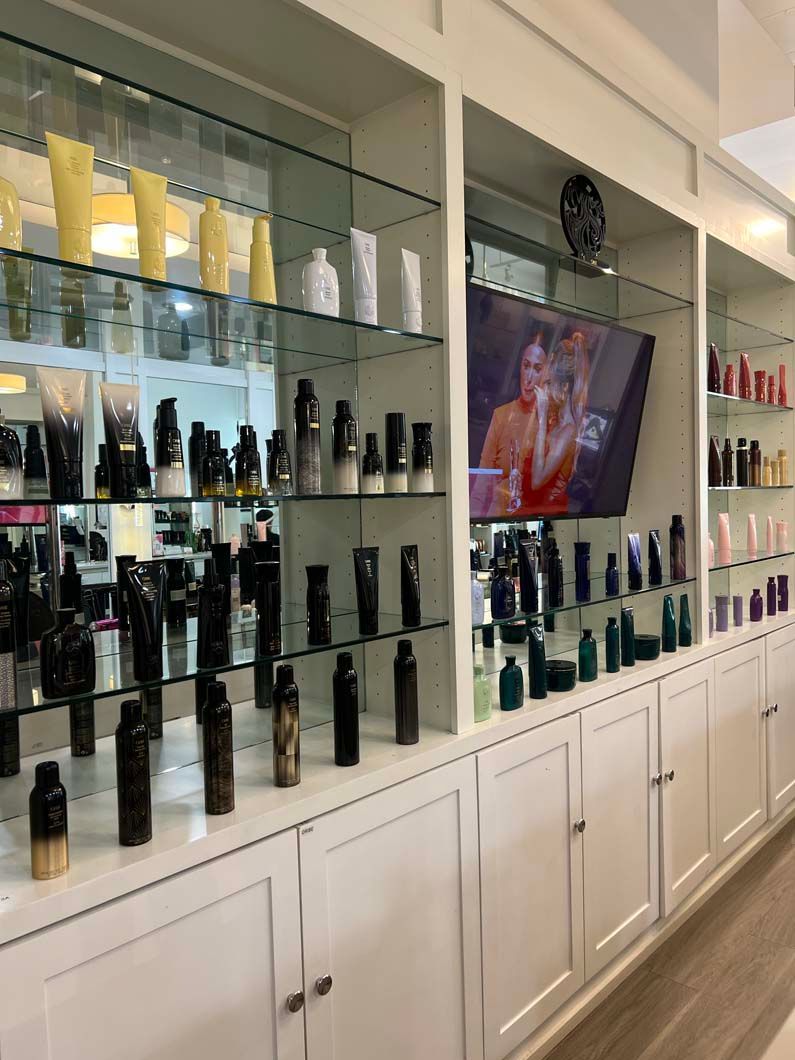 Shelves displaying hair products in a salon. A TV screen shows a person. White cabinetry lines the walls.