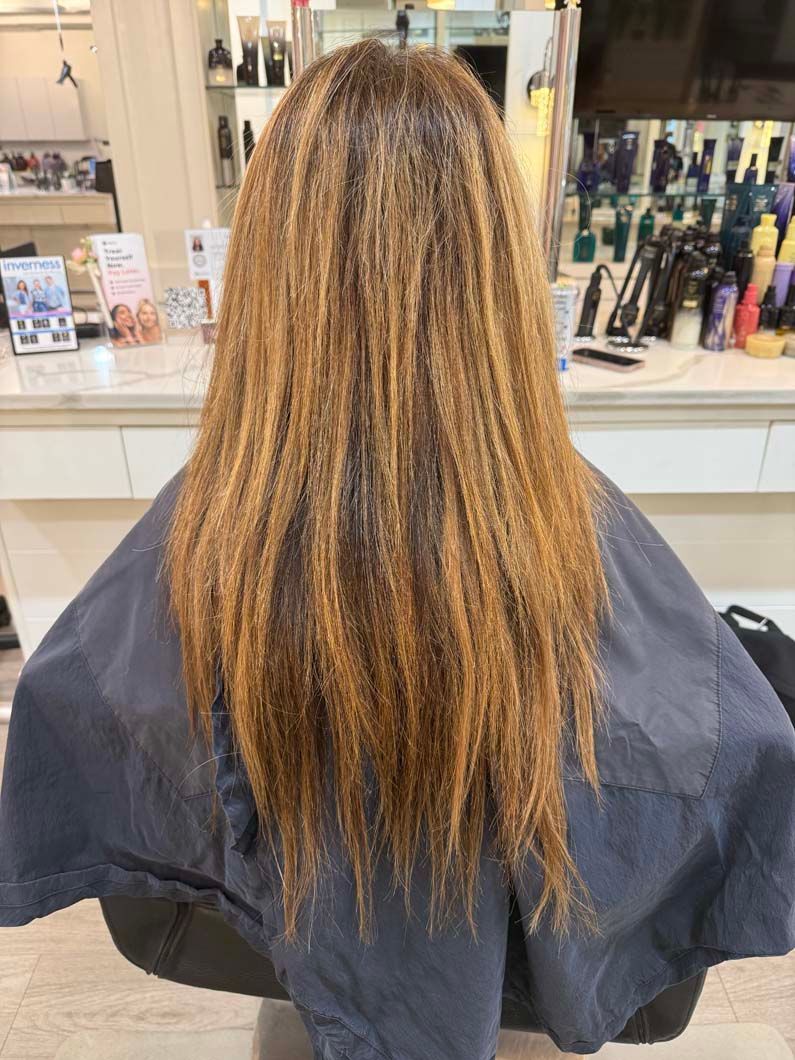 Woman with long, layered brown hair, some streaks lighter. Sitting in salon chair with cape on.