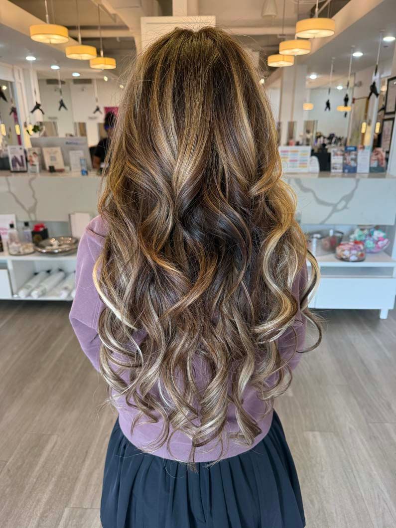Woman with long, wavy brown hair with blonde highlights. Standing in a salon.