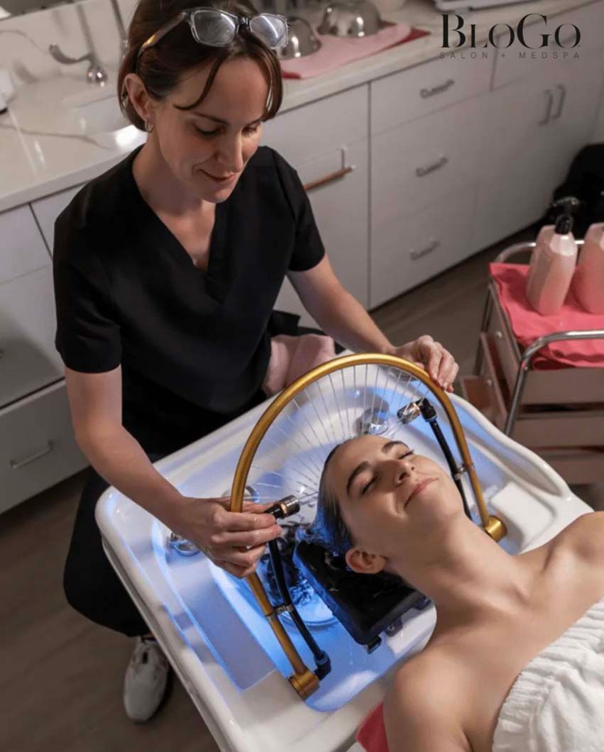 Woman receiving treatment in a spa setting; technician holds device over her head, blue light shines.