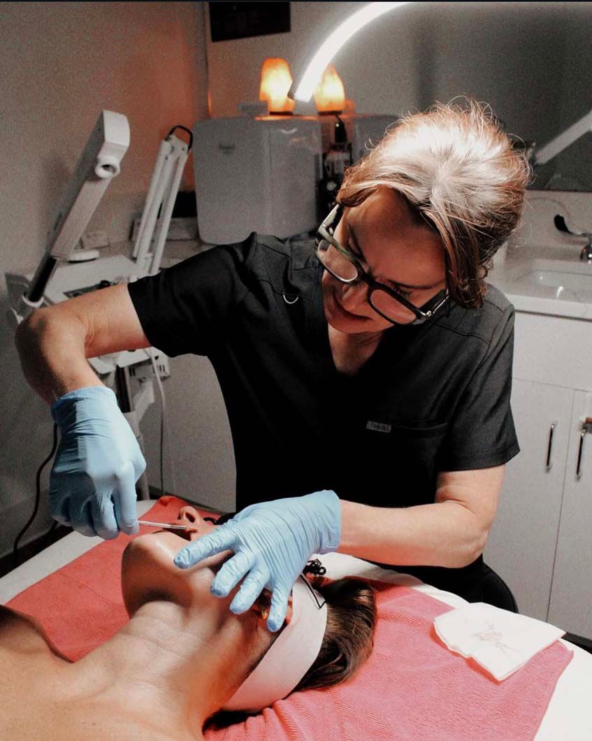 Aesthetician in blue gloves performing facial treatment on a client with a towel; medical setting.