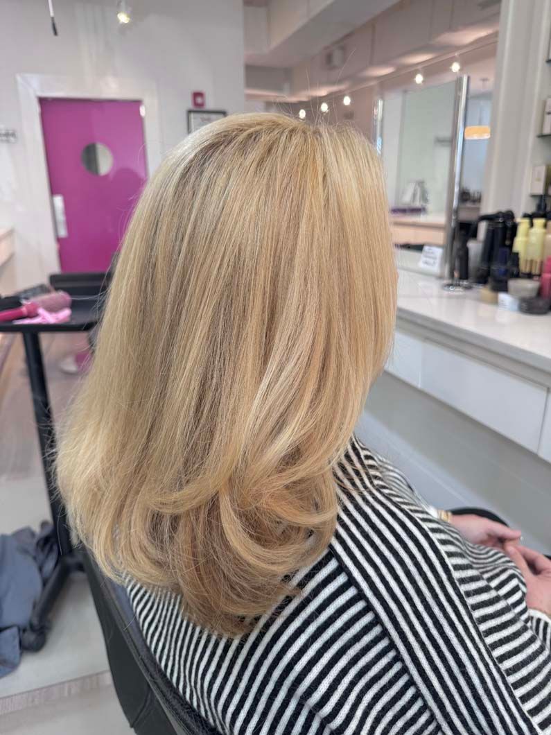Woman with layered blonde hair sitting in a salon chair; wearing a striped shirt.