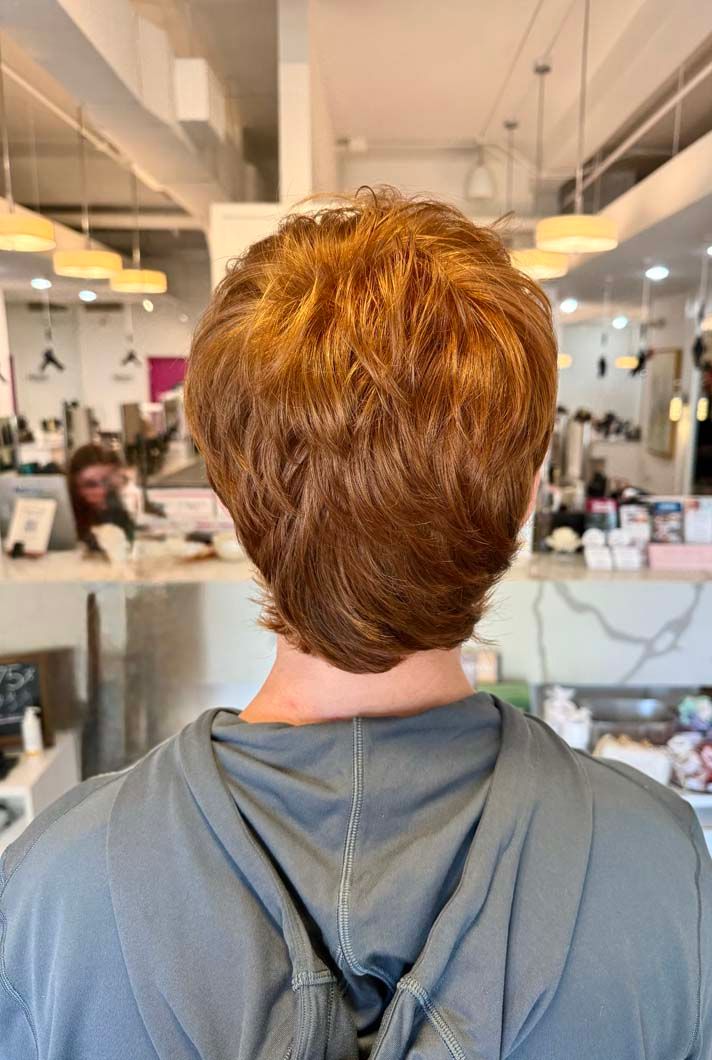 Back view of a person with short, reddish-brown hair in a salon; they wear a grey hoodie.