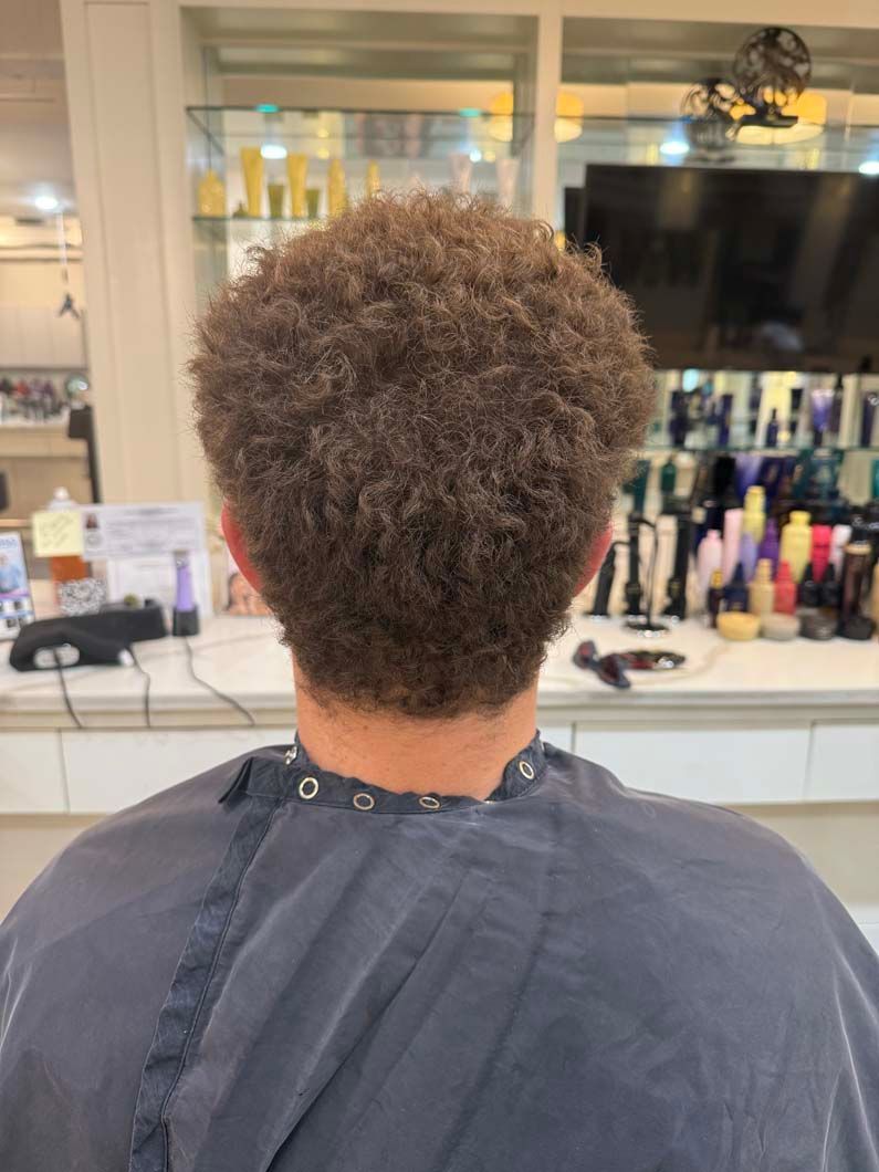 Person in a salon chair, facing away. Brown curly hair, black salon cape.