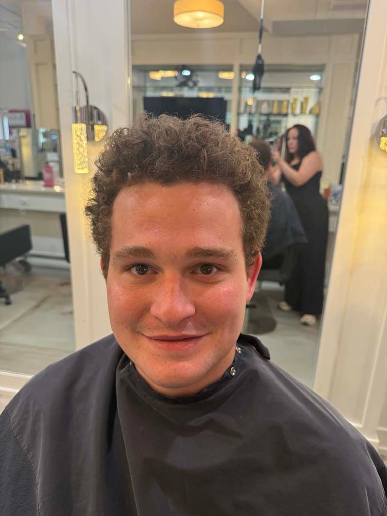 Man with curly brown hair in a salon. He is smiling while wearing a black cape.
