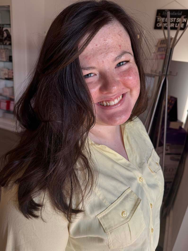 Woman with brown hair and a light yellow shirt smiles indoors.
