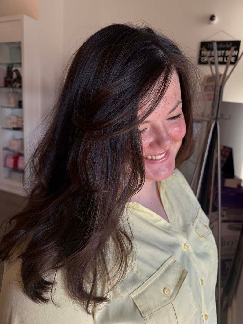 Woman with long dark hair, smiling, wearing a light yellow shirt, indoors.