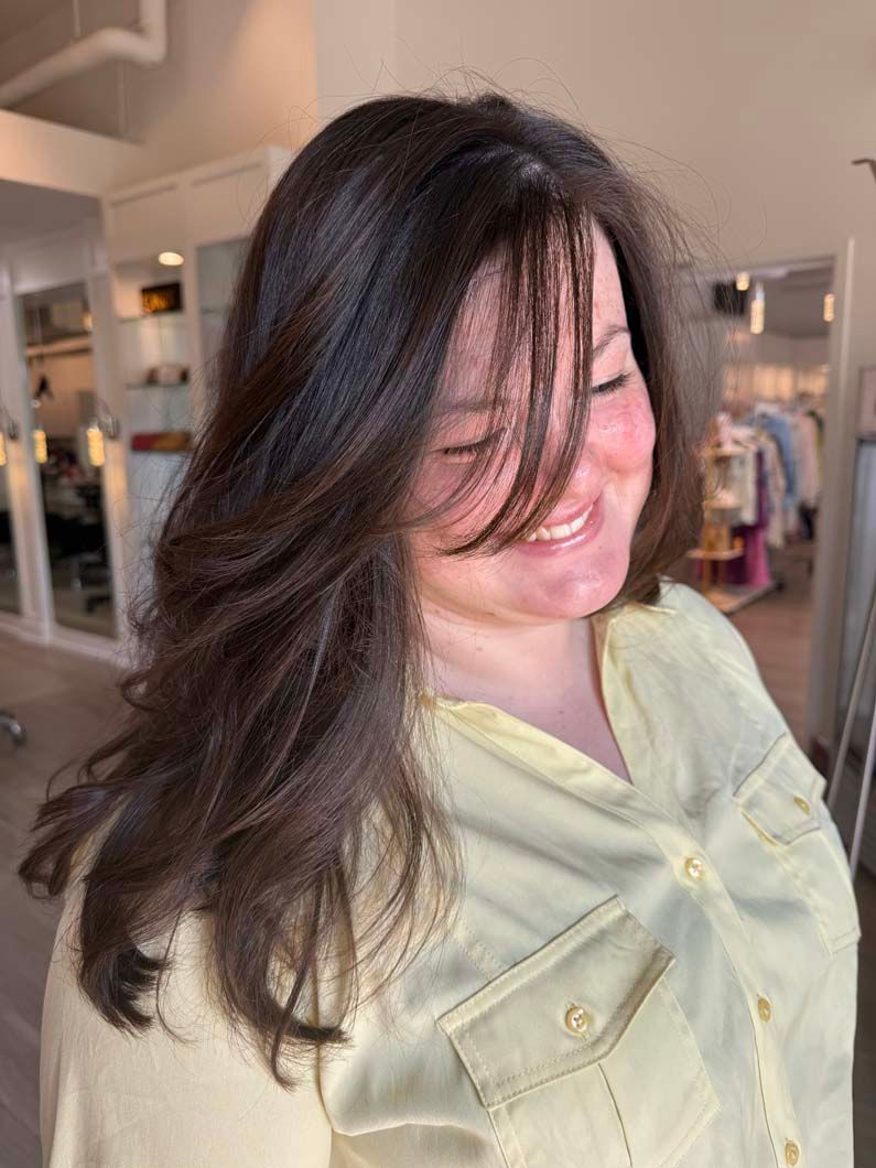 Woman with dark hair smiles, wearing a yellow shirt, in a salon. Hair frames her face.
