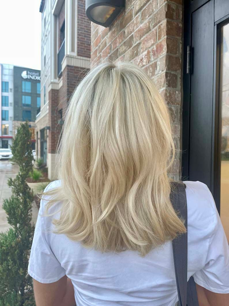 Blonde shoulder-length hair styled with waves, woman in white shirt, standing by brick building.