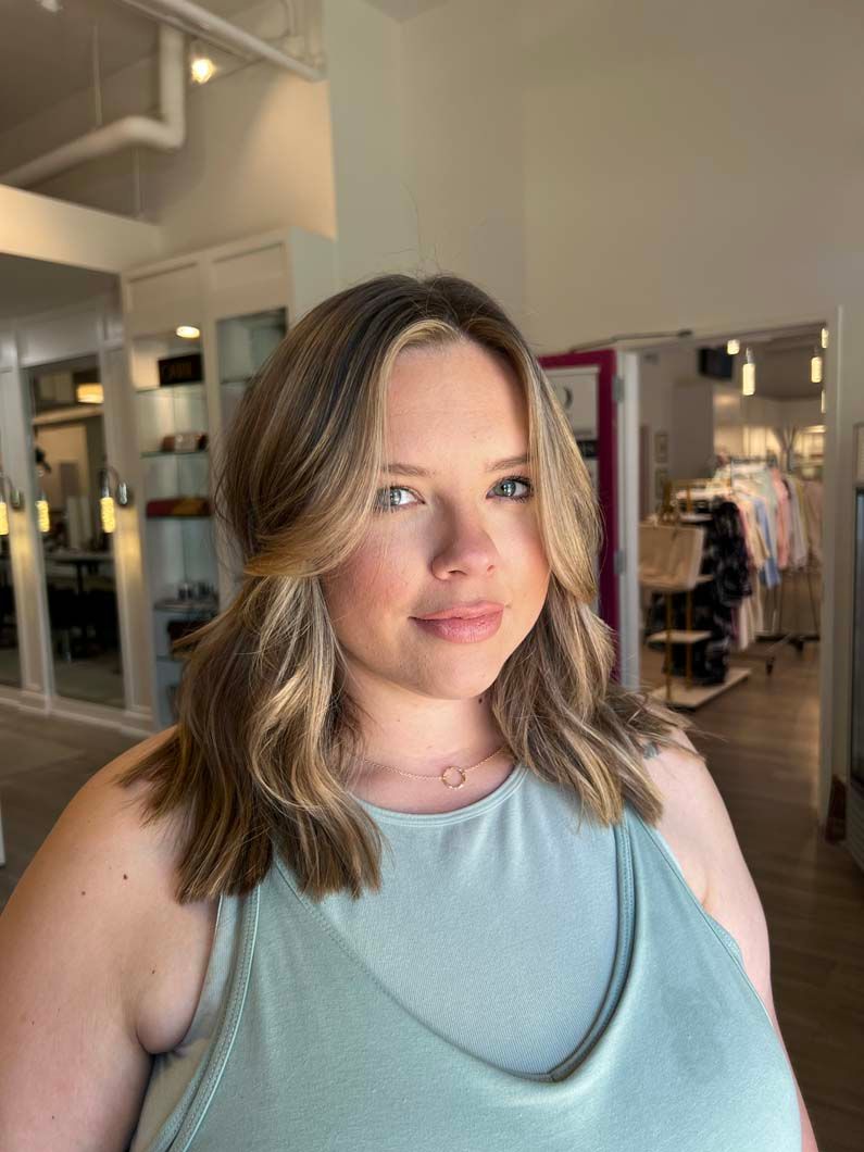 Woman with wavy, highlighted hair in a salon, wearing a light blue tank top and delicate necklace.