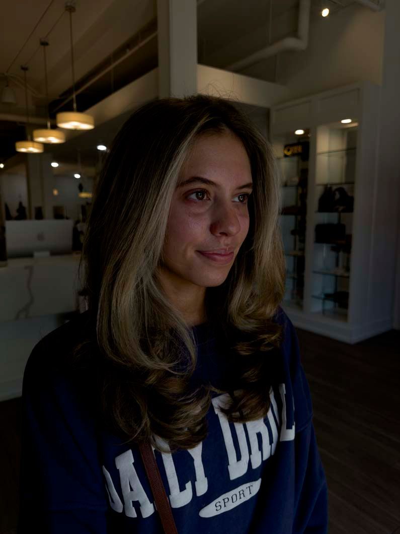 Woman with wavy hair, wearing a blue sweatshirt, in a brightly lit salon.