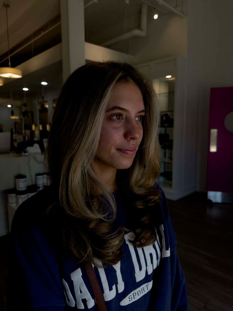 Woman with layered brown hair and blonde highlights, in a salon. Wearing a navy sweatshirt.