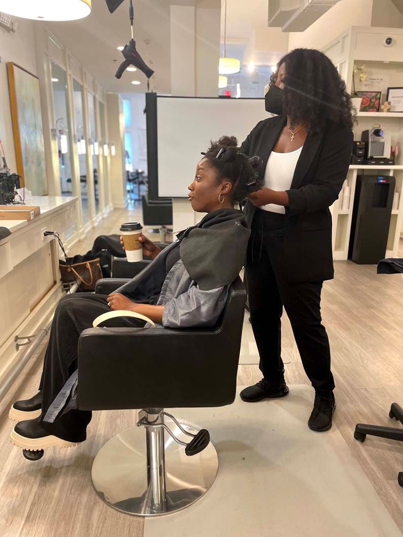 Woman getting her hair styled at a salon. Stylist wearing a mask and black suit is holding the woman's hair.